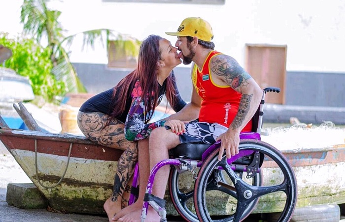 Una mujer con tatuajes y en traje de baño, está sentada en una lancha que está en tierra y besa apasionadamente a un hombre en silla de ruedas. 