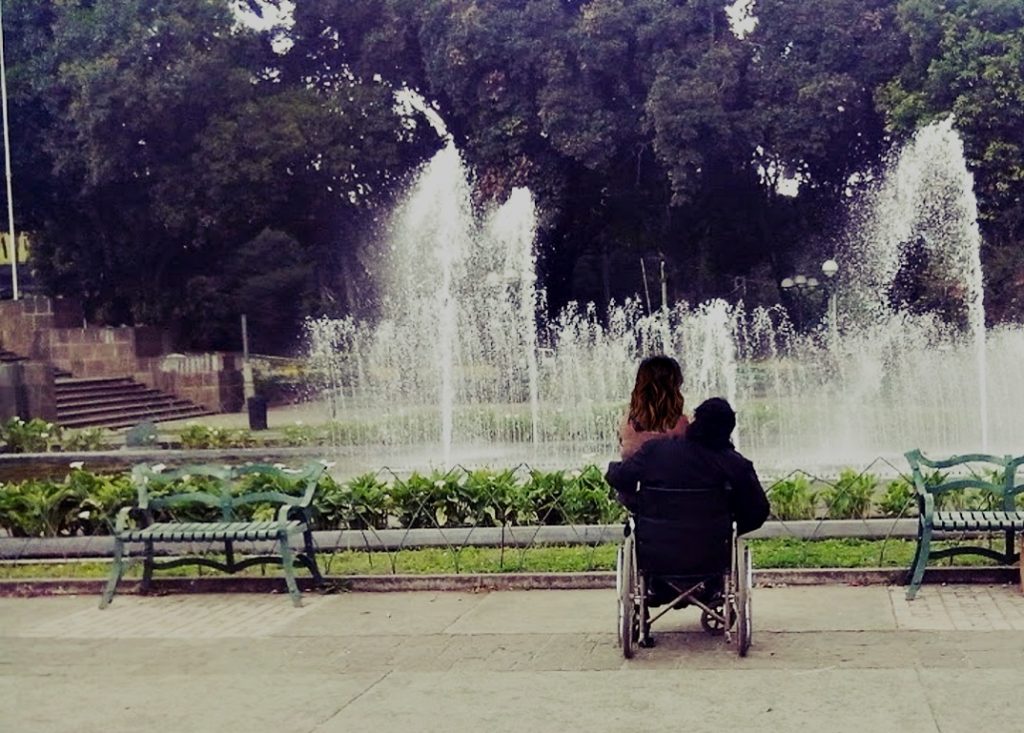 En un parque, frente a una fuente, se vea Byron Pernilla de espaldas en su silla de ruedas, en sus piernas una mujer de pelo largo castaño. 
