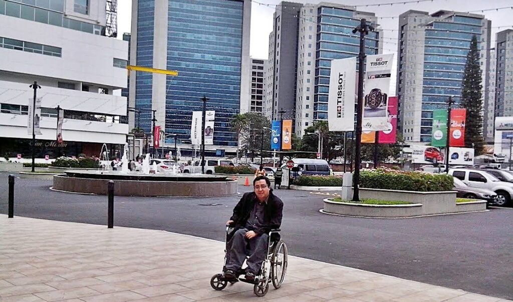 Fotografía de Byron Pernilla sentado en silla de ruedas vestido con chaqueta negra, se observa en una plaza comercial, al fondo se ven edificios.