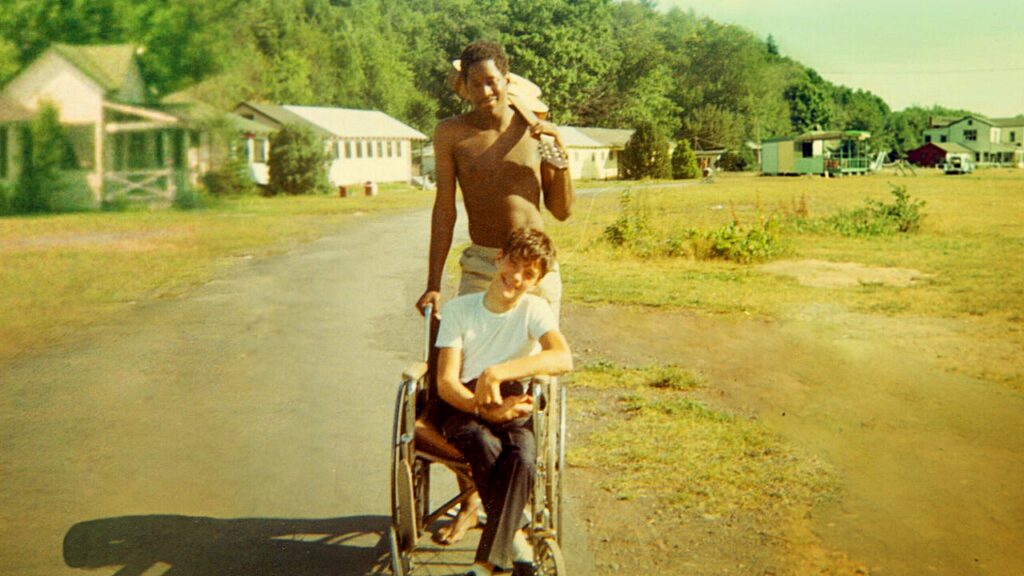 Fotografía antigua, se observa en campo con algunas galeras a la distancia, al frente un chico en silla de ruedas y atrás de él un joven afroamericano sin camisa y una guitarra al hombro.