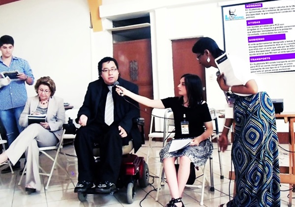 Byron Pernilla, de traje oscuro y corbata, habla por un micrófono que le sostiene una chica que está sentada a su lado, atrás algunas personas toman nota de la capacitación.