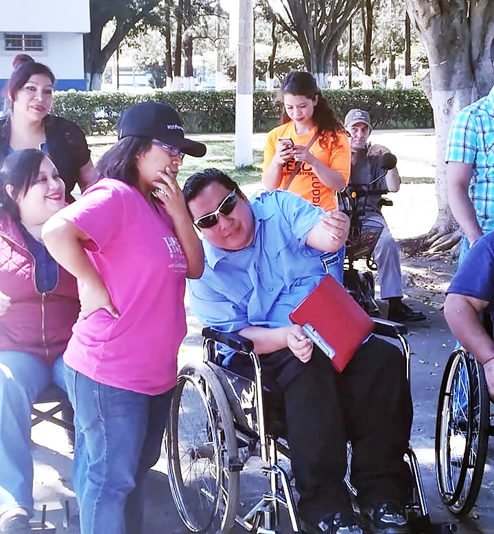 Byron Pernilla, en su silla de ruedas y con un folder en las piernas, conversa con una voluntaria durante un evento, atrás, se ven más personas con y sin discapacidad.