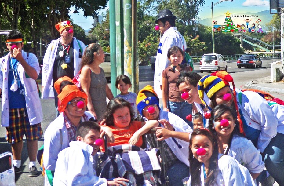 Una niña en silla de ruedas está rodeada por varios voluntarios con batas blancas y nariz roja de payaso. Está el logo del Paseo Navideño ASODISPRO.