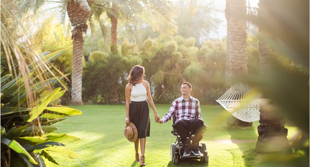 Una pareja camina de la mano en un jardin en una tarde soleada, él en una silla de ruedas eléctrica y ella caminando