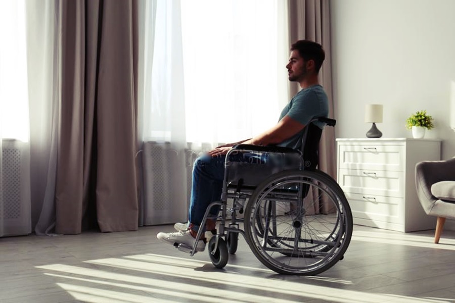 Una persona con discapacidad, en su silla de ruedas en una habitación semi-oscura con ventanas soleadas.