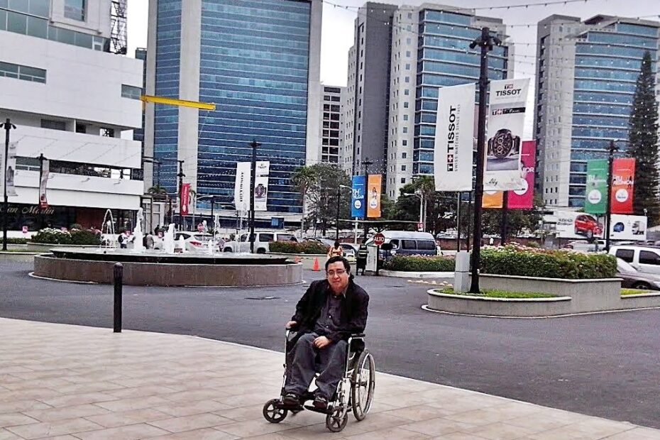 Fotografía de Byron Pernilla sentado en silla de ruedas vestido con chaqueta negra, se observa en una plaza comercial, al fondo se ven edificios.