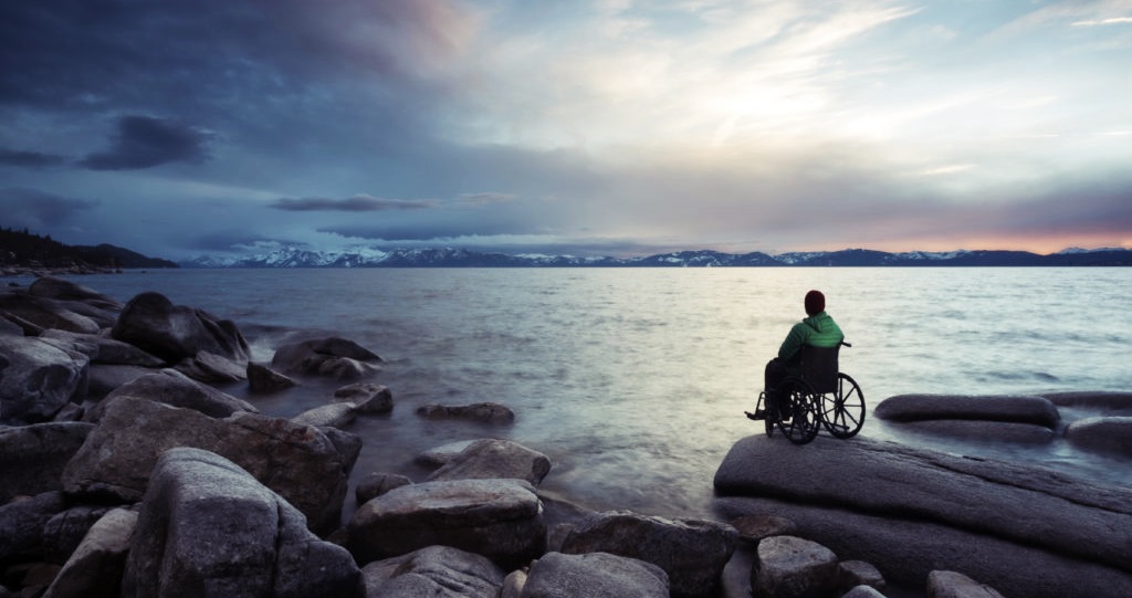 En un paisaje de rocas, mar y nubosidad, una persona con discapacidad en silla de ruedas está sobre una roca, como observando el horizonte