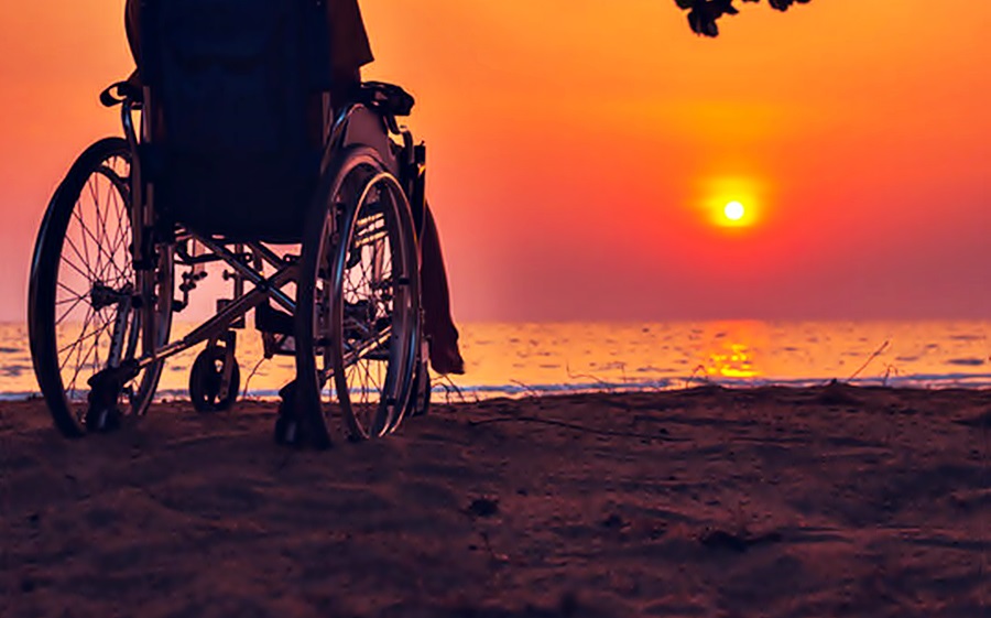 En un atardecer frente al mar, se ve una silla de ruedas y al fondo un sol amarillento con un contorno anaranjado.