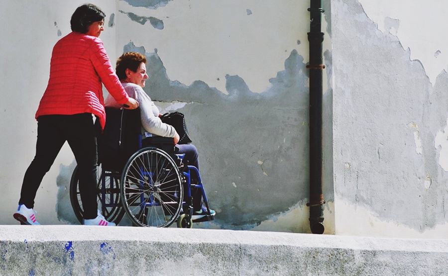 En la acera de una calle, se observa a una mujer empujar la silla de ruedas de otra mujer.