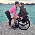 Jessica Arevalo y Garrett Greer, están en un muelle, ella de pie con ropa deportiva y el en su silla de ruedas, al fondo se ve un pequeño oleaje, en un día nublado.
