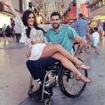 En un centro comercial, Jessica Arevalo y Garrett Greer se toman una fotografía con gente caminando a sus espaldas. Ella viste una minifalda y una blusa de encaje pequeña, está sentada en las piernas de él, quien está en su silla de ruedas.