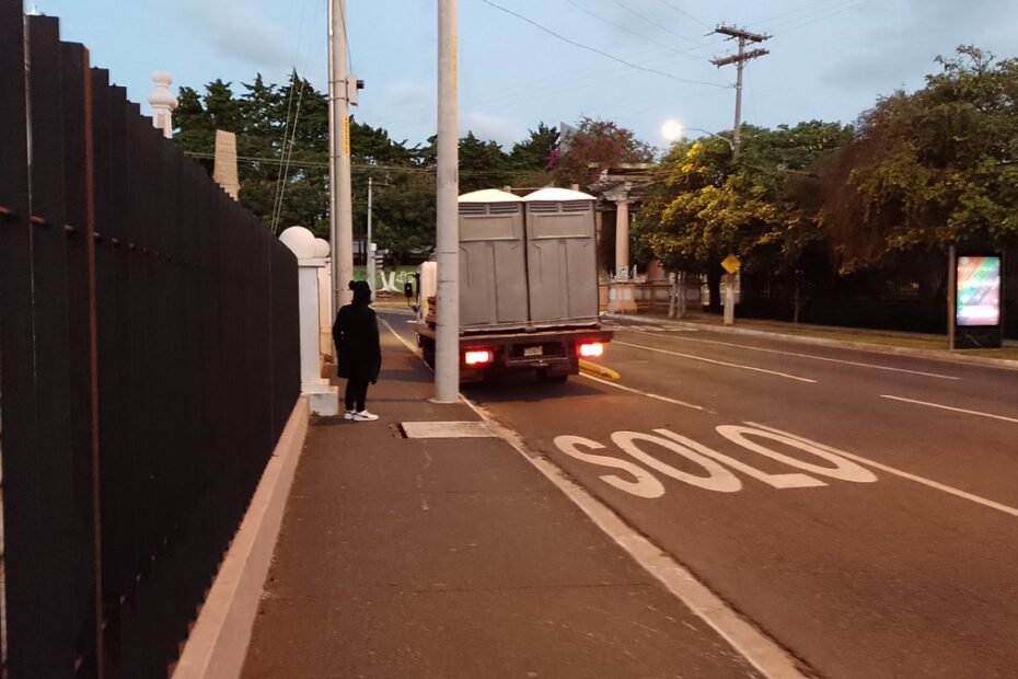 De madrugada, en una calle desierta una voluntaria está a la par de un camión con baños portátiles